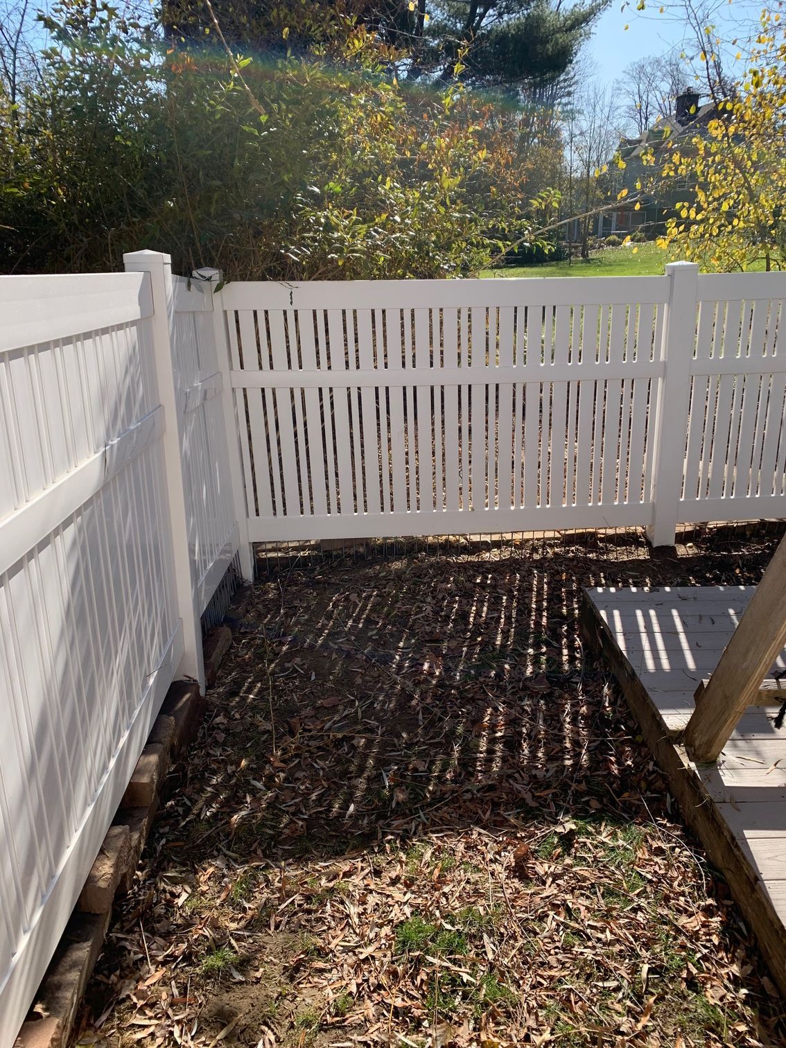 White vinyl fence with an opening, partially enclosing a yard with brown leaves and sunlight shadows.