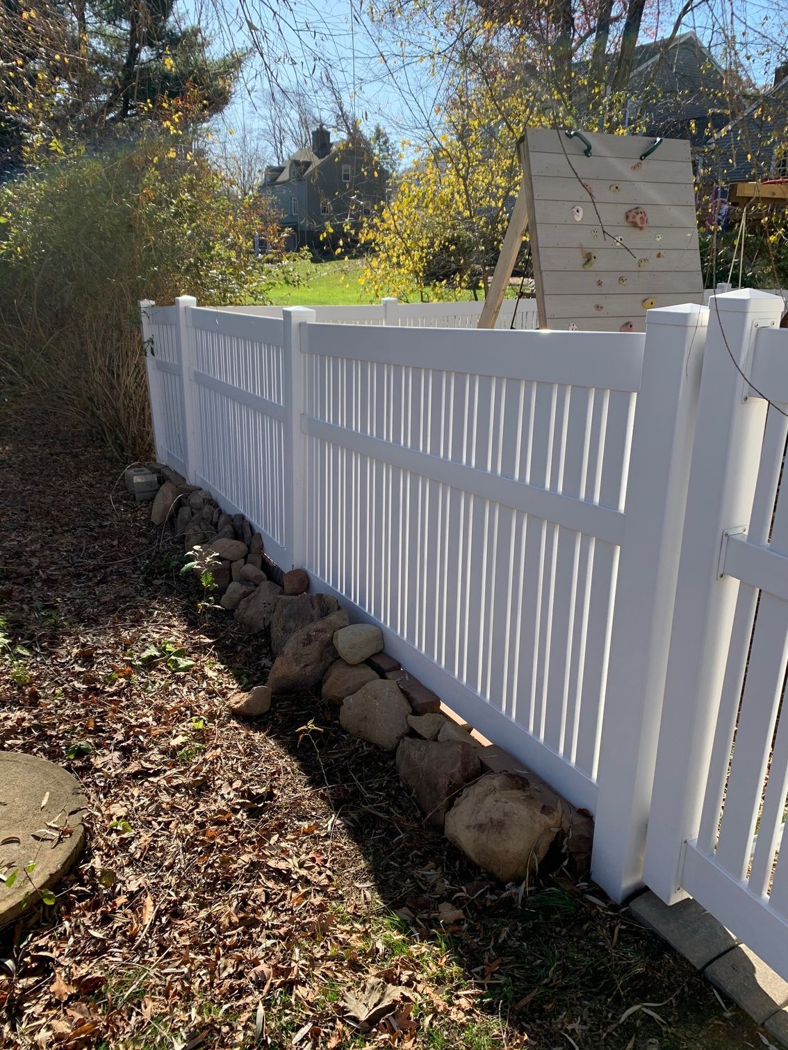 White vinyl fence bordering a yard with a row of rocks at the base. A wooden target is visible in the background.