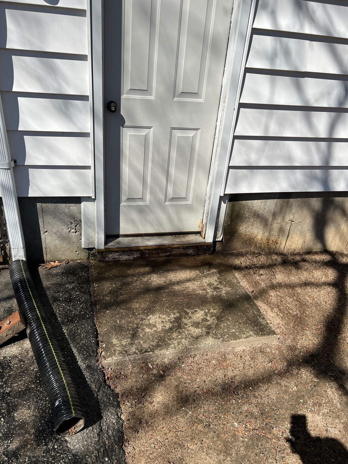 White door leaning, concrete patio uneven with foundation. White siding and gutter visible.