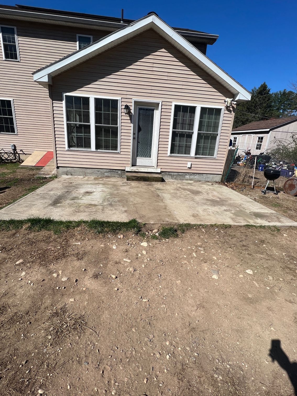 Backyard with a concrete patio, beige house, and a door. Dirt ground in front with some grass.