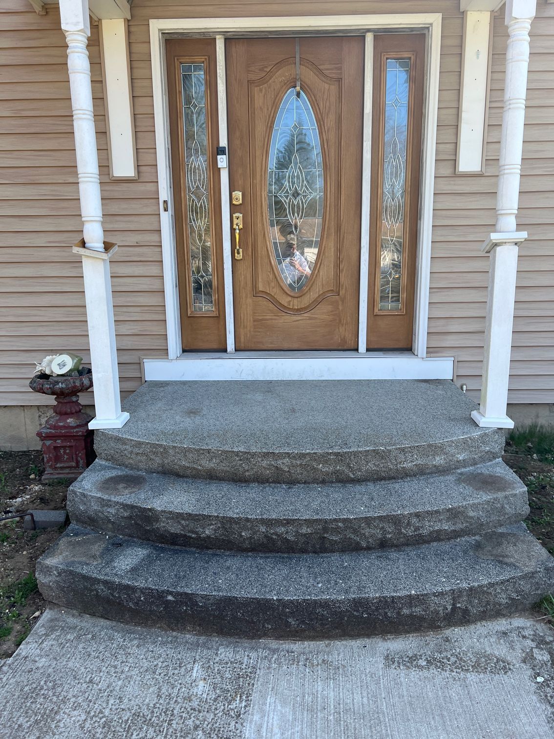 Three concrete steps leading up to a front door with glass panels. A porch with white pillars and a vase is to the left.