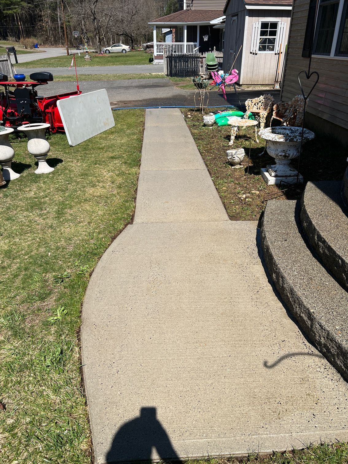 A concrete walkway leads to steps and a house with various items on the lawn, including a red cart and garden ornaments.
