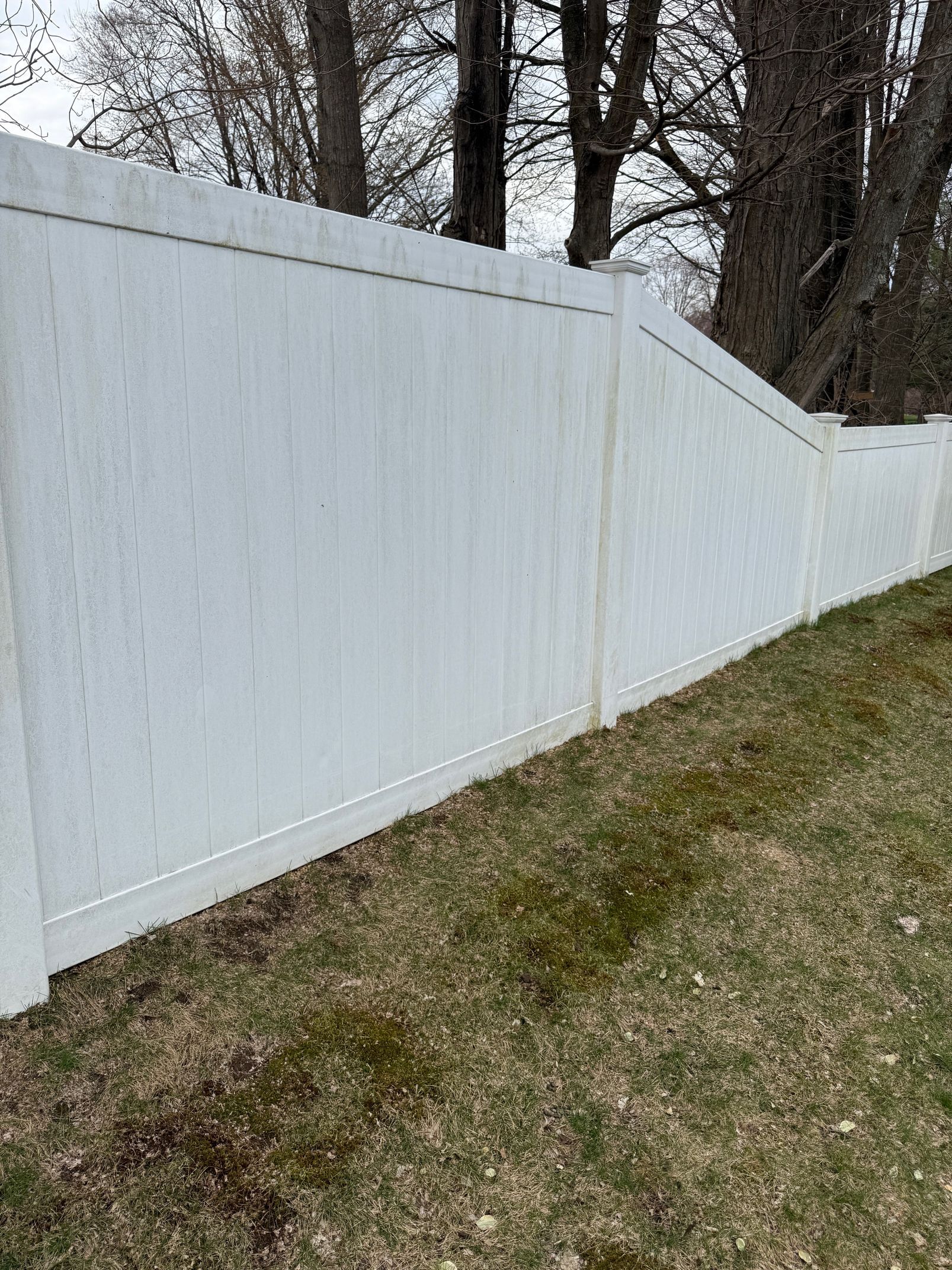White vinyl fence running along a grassy yard, with trees in the background. The fence has some discoloration.