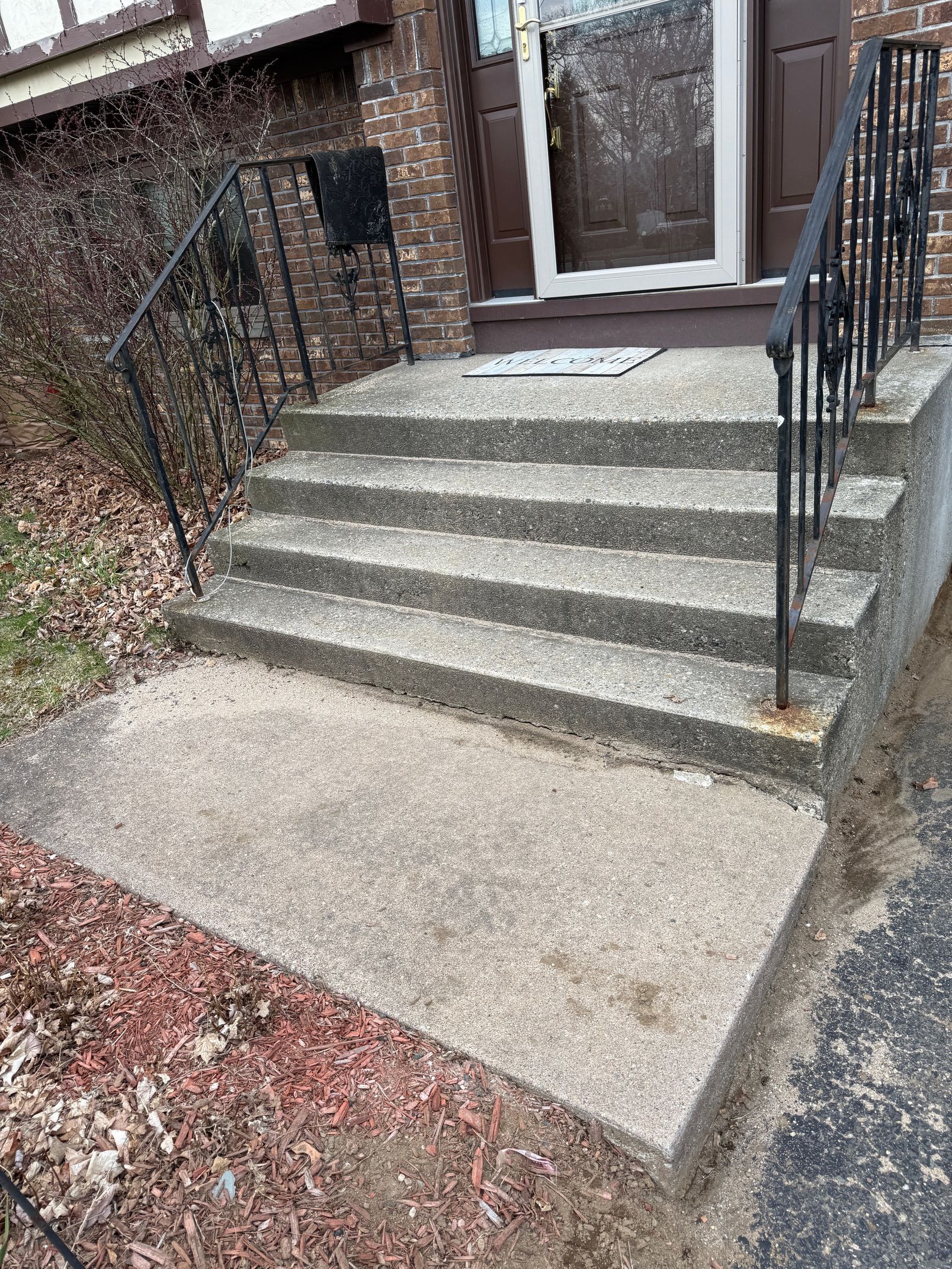 Concrete steps leading up to a brown door, with a wrought-iron railing on the right and left.