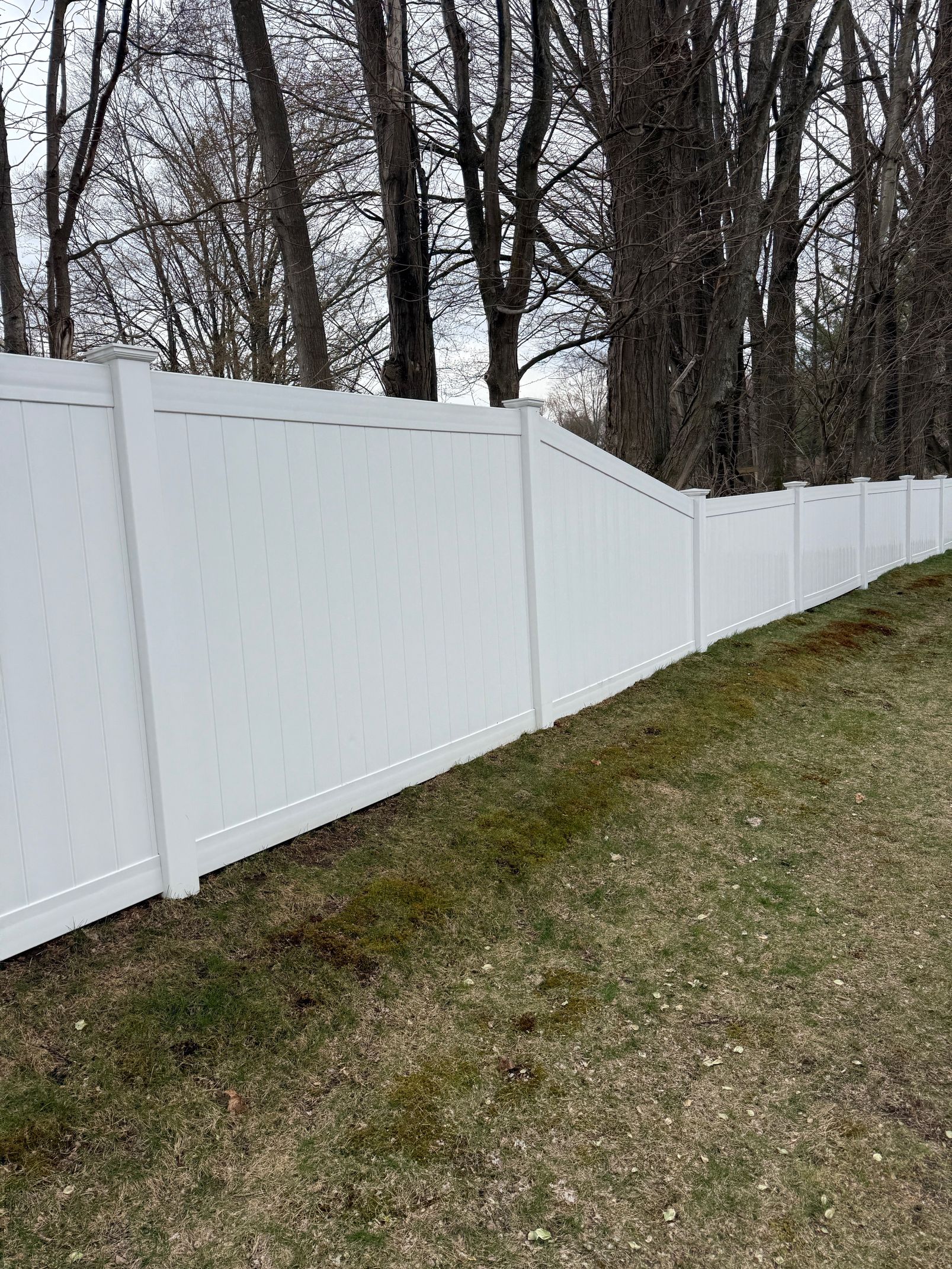 White vinyl fence along a grassy slope, with bare trees in the background.