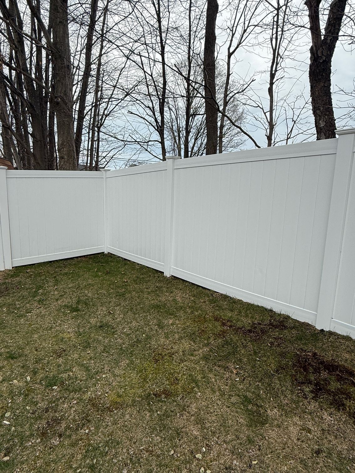 White vinyl fence in a yard with brown grass and bare trees in the background.