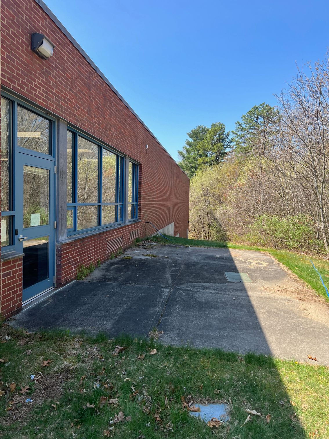 Brick building with windows and door; a concrete path runs alongside grass and greenery on a sunny day.