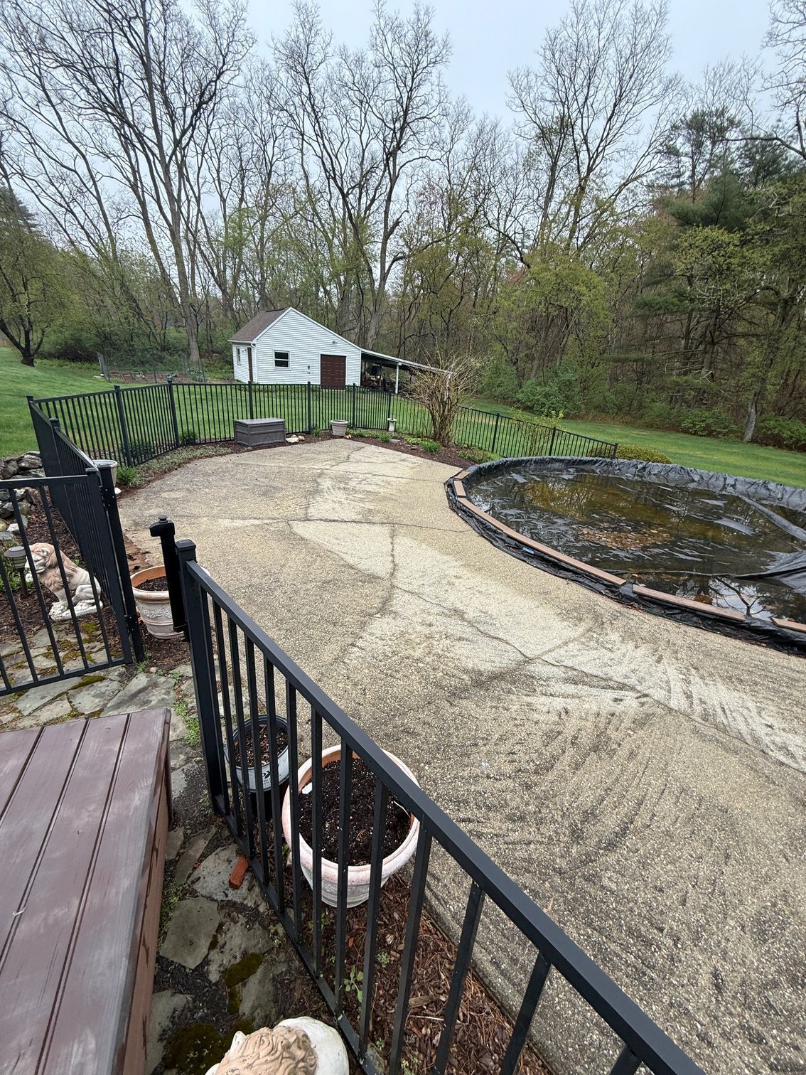 A backyard scene with a pool covered in a tarp, a cracked concrete patio, and a small shed, surrounded by a fence and trees.