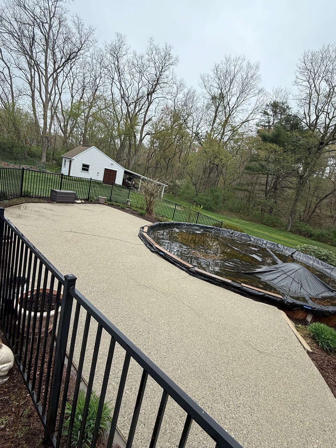A backyard with a partially covered pool, gravel path, and a small white shed surrounded by trees.