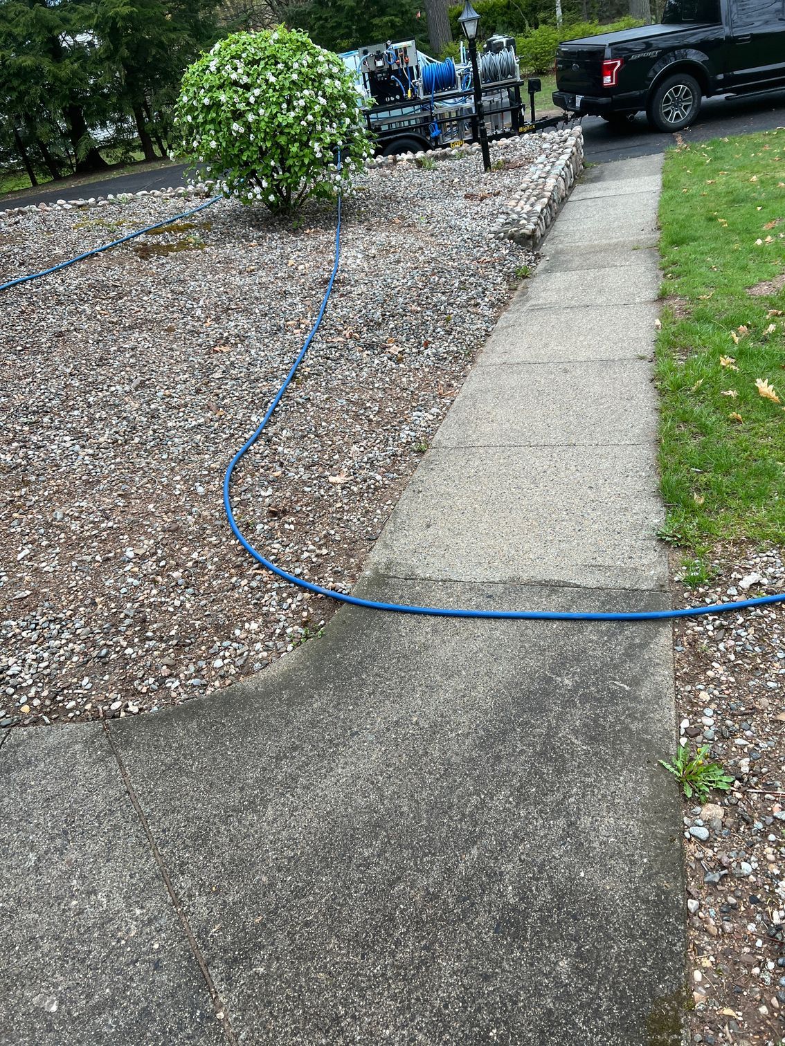 A concrete sidewalk with a blue hose running beside it, next to a rock garden and a truck.
