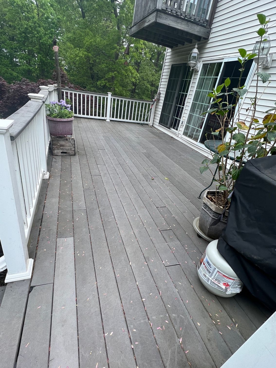 Wooden deck with white railing next to house with glass doors. Flowers in pots, propane tank visible.