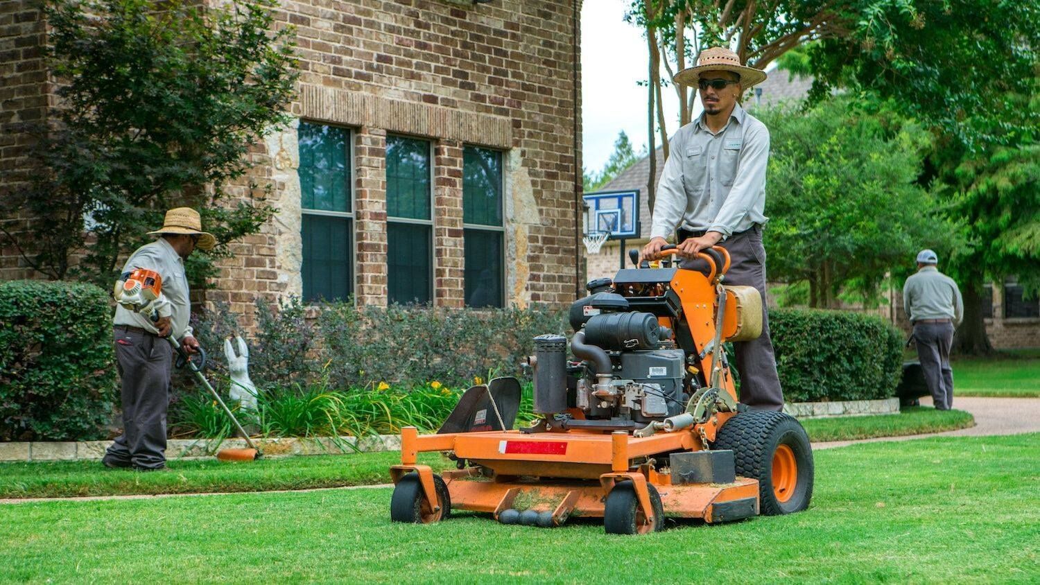 Landscapers mowing a lawn with orange riding mowers beside a brick building