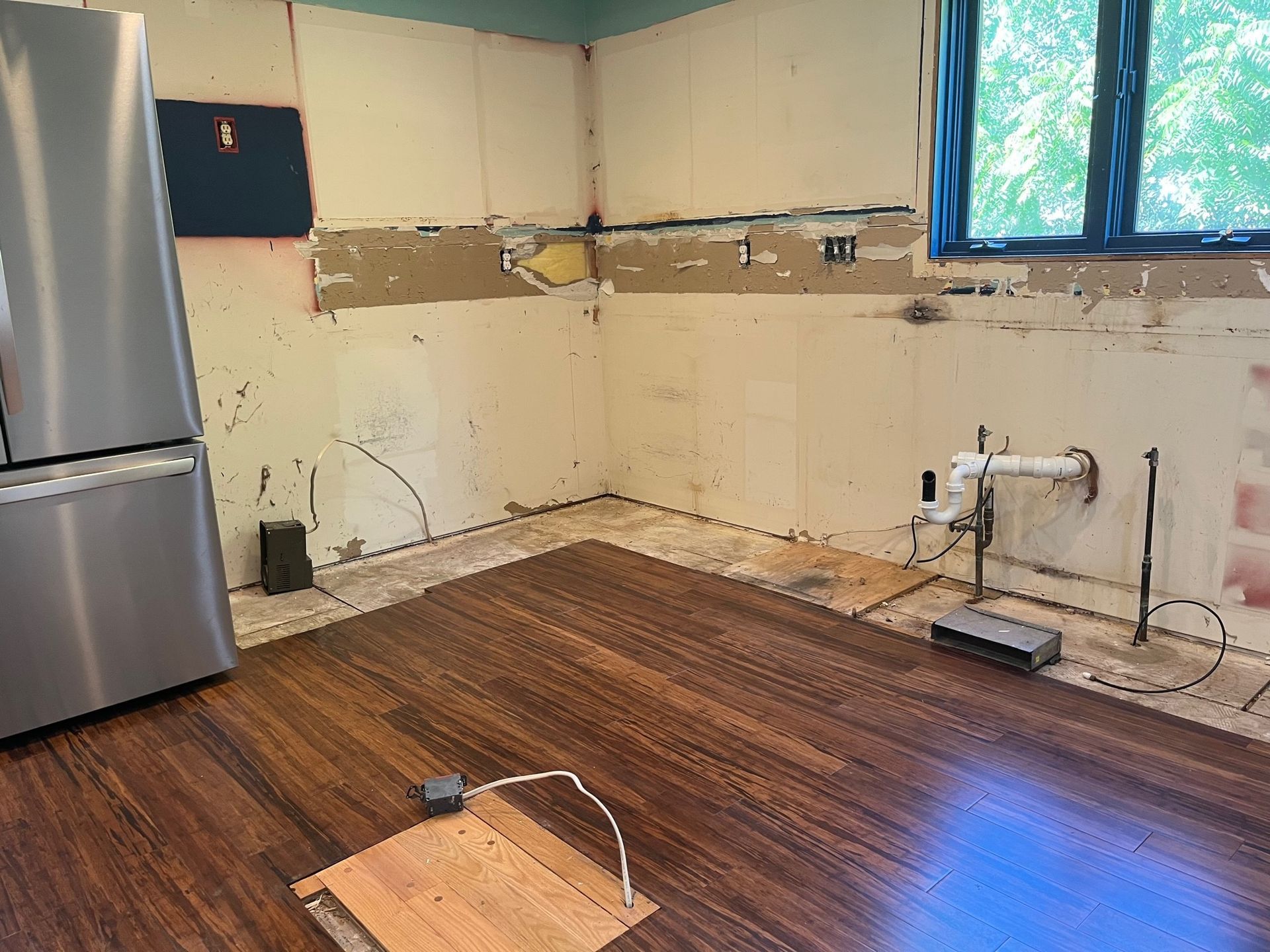 Kitchen renovation: bare walls, exposed plumbing, new wood flooring, stainless steel fridge.