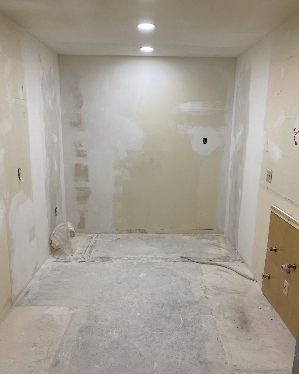 Empty room under construction with recessed lighting. Bare walls and floor, drywall patching visible.
