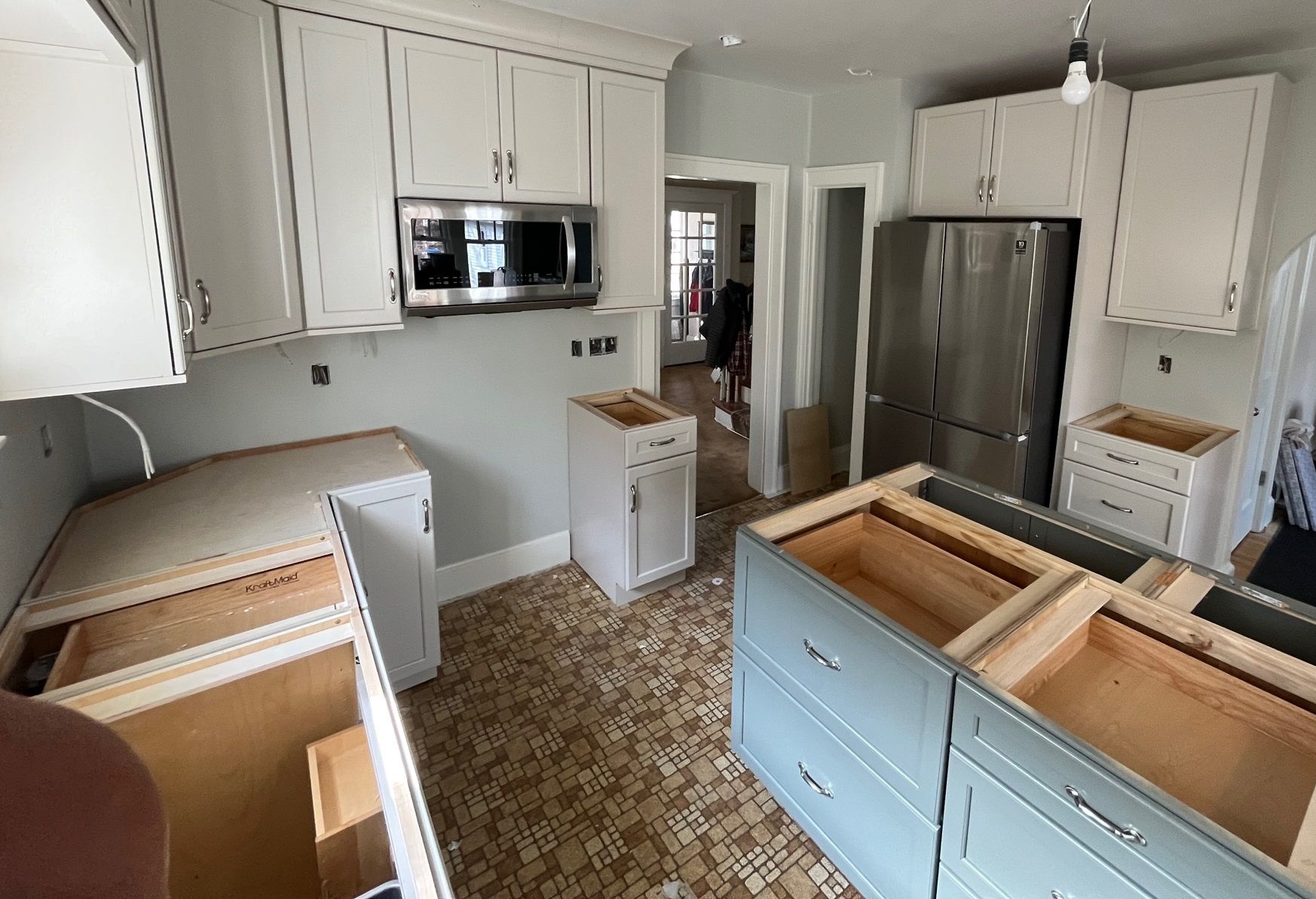 Kitchen under renovation with white cabinets, stainless steel appliances, & blue island; flooring is brown and yellow tiles.