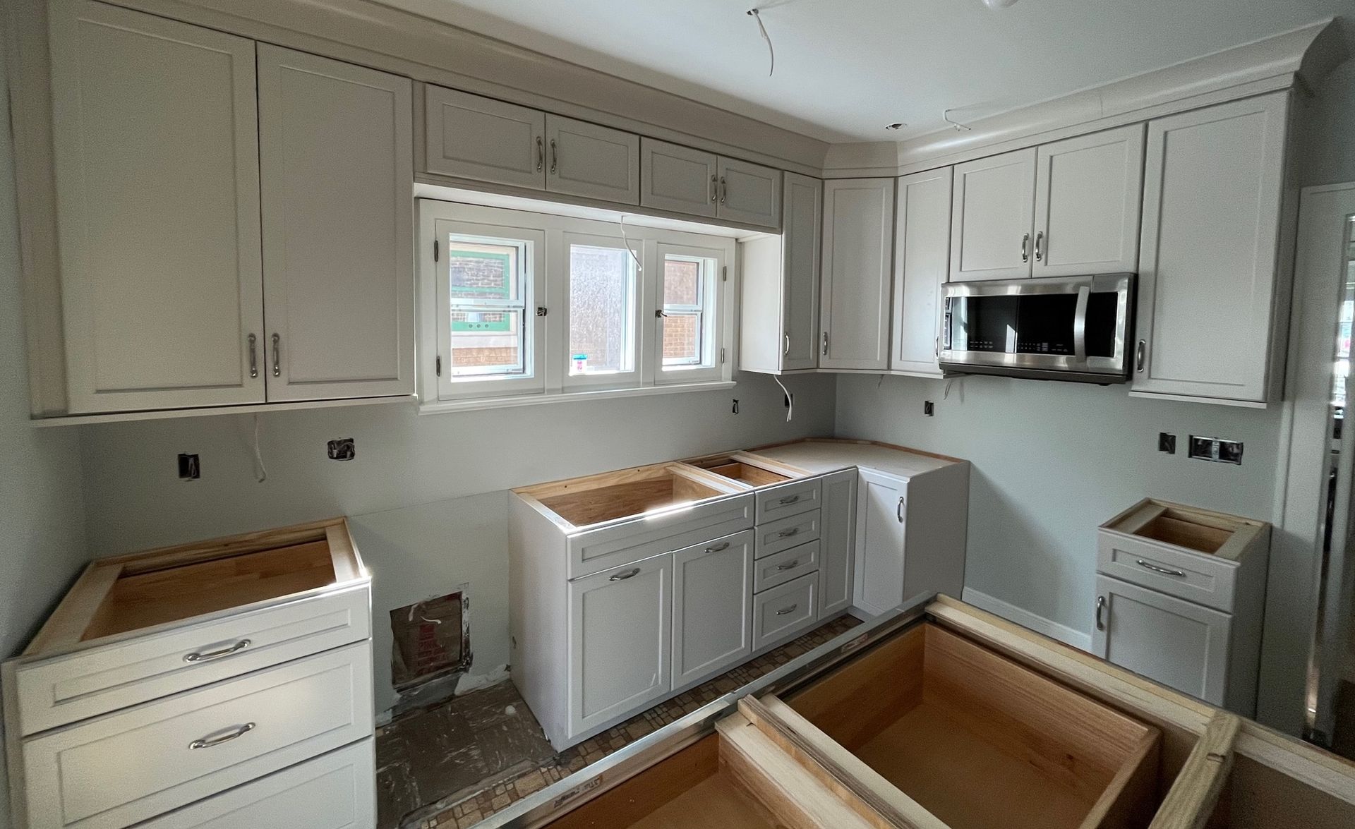Kitchen cabinets being installed; white upper and lower cabinets, stainless steel microwave, unfinished countertops.