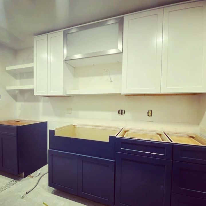 Kitchen under construction with blue and white cabinets, stainless steel range hood, and floating shelves.