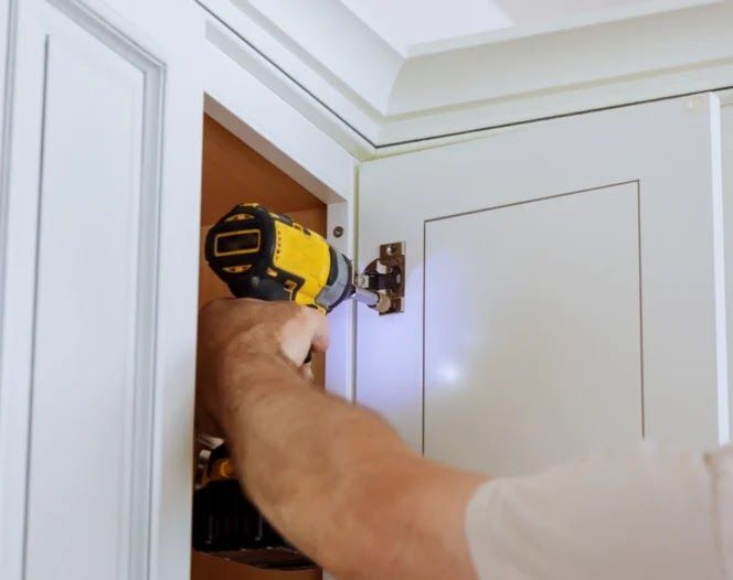 Person using a yellow drill to install a cabinet hinge on a white cabinet.