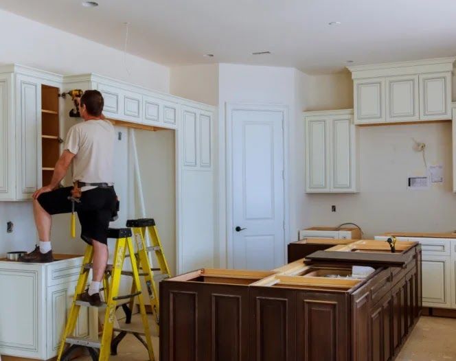 A person installing white kitchen cabinets on a yellow ladder; dark brown island in the center.