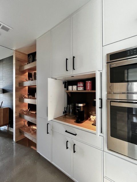 White built-in kitchen cabinetry with a coffee station. Features an oven, wooden shelves, and a coffee maker.