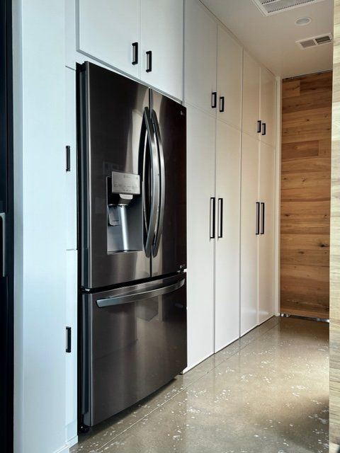 Stainless steel refrigerator flanked by white cabinets and a wooden wall, on a concrete floor.