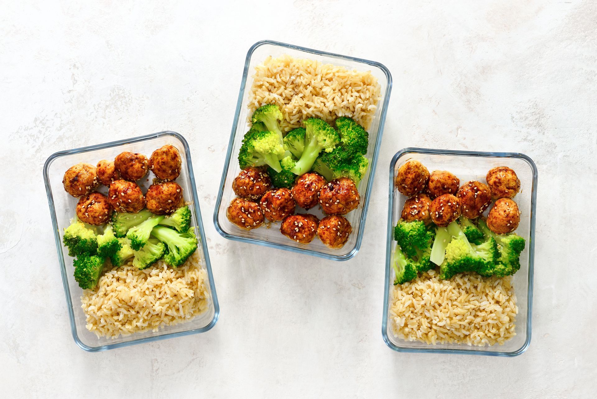 Three glass containers filled with meatballs , broccoli and rice.