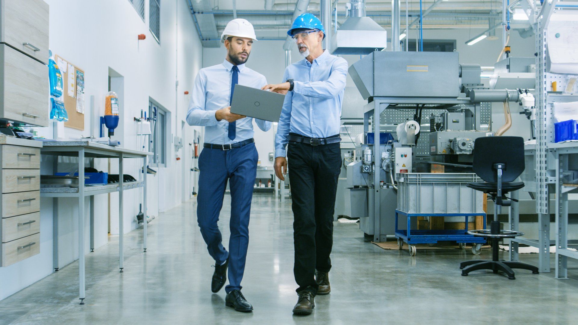 Two men are walking in a factory and looking at a tablet.