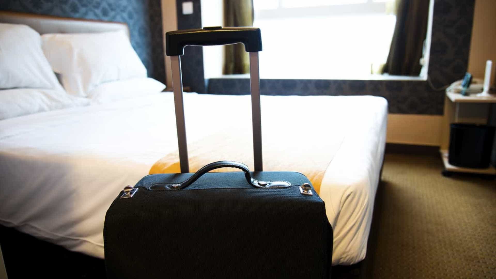 Quarto de hotel com mala de viagem azul em primeiro plano. Papel de parede azul, roupa de cama e almofadas brancas
