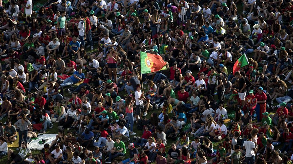Multidão de jovens sentados na relva. No centro da imagem, um rapaz segura a bandeira de Portugal
