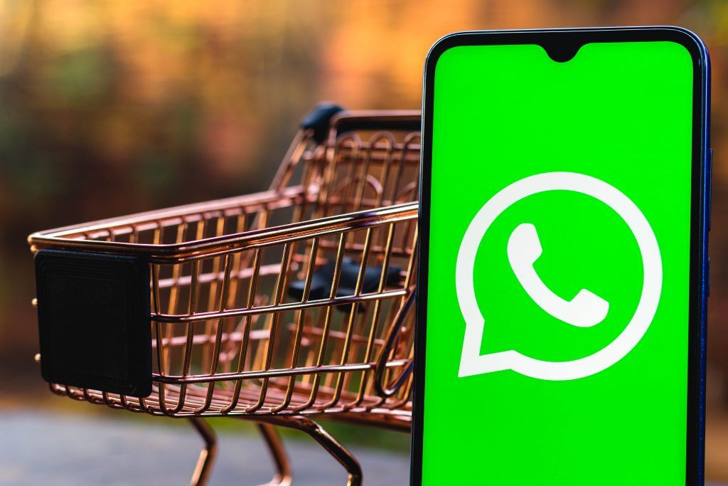 Ecrã de telemóvel com o símbolo do WhatsApp. Carrinho de compras metálico no fundo.