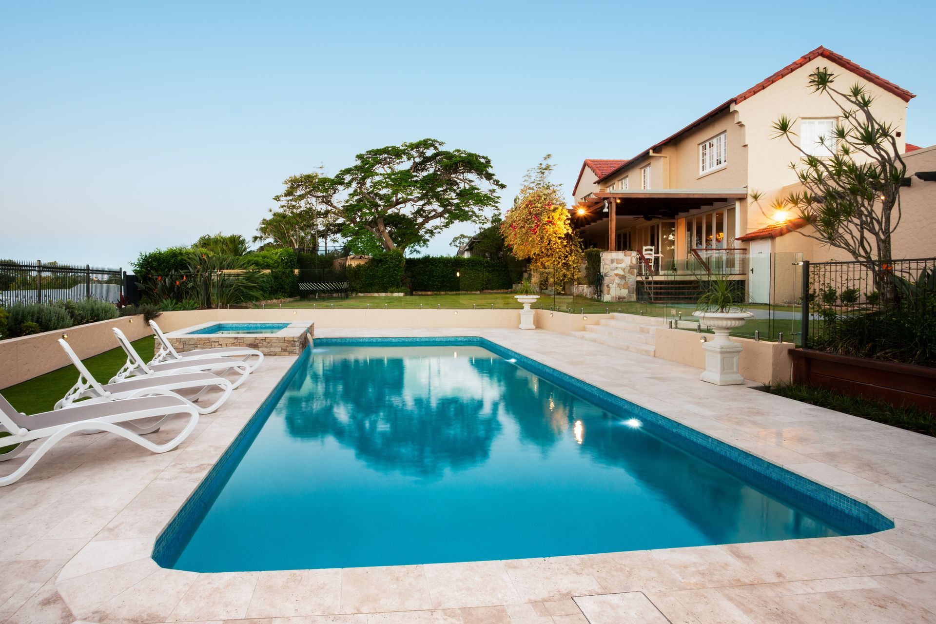 A rectangular swimming pool with turquoise water sits on a stone patio beside a large house at dusk.