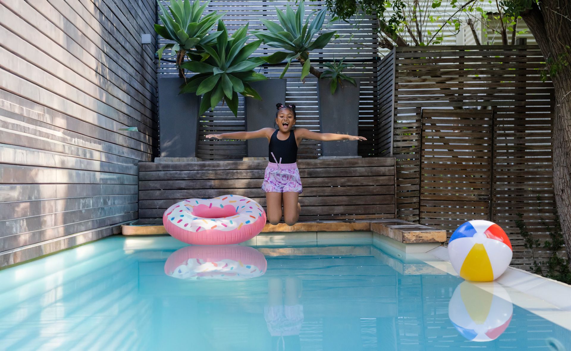 A girl wearing swimwear kneeling on a bench at a backyard pool, reaching towards a doughnut float. A girl wearing swimwear kneeling on a bench at a backyard pool, reaching towards a doughnut float.