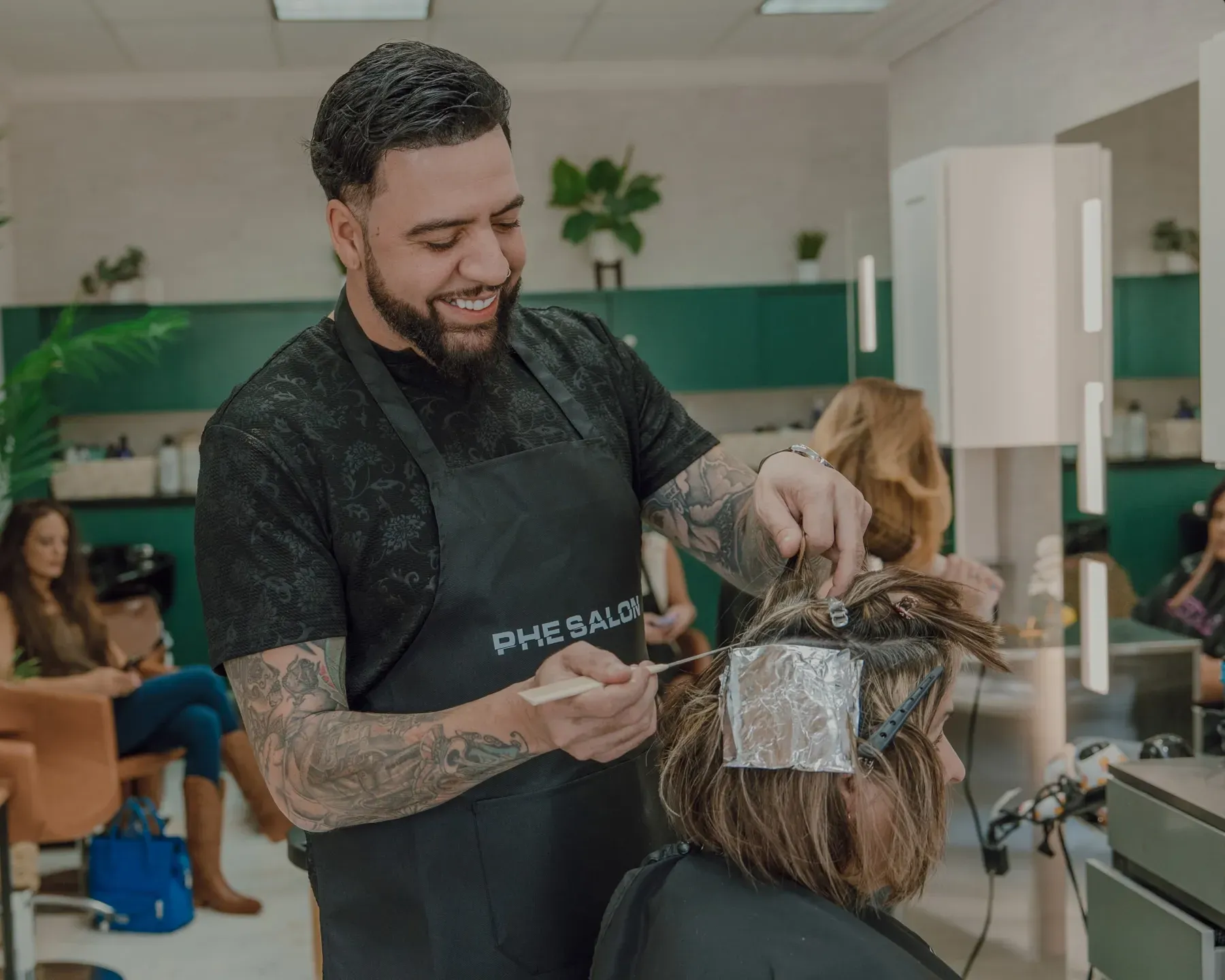 Hair stylist applying foil highlights in a salon, smiling. Green wall backdrop with clients.
