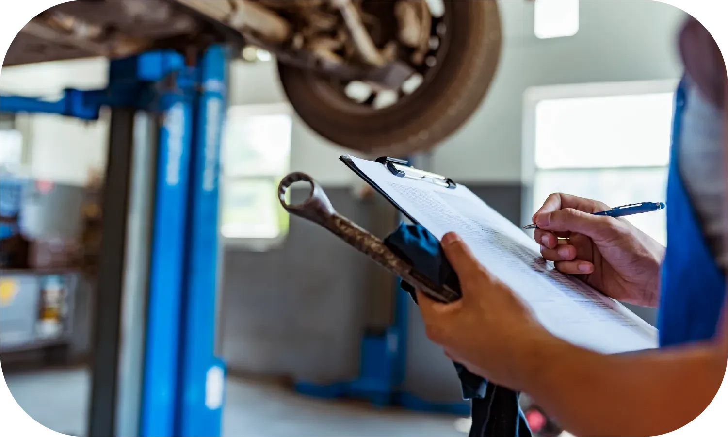 A mechanic is writing on a clipboard in front of a car on a lift.
