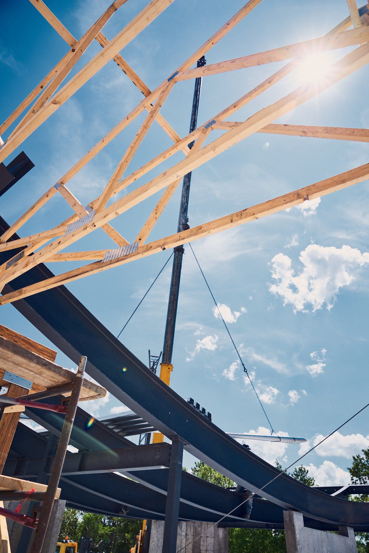 Chantier de construction : Grue soulevant une poutre d'acier pour la poser sur une charpente de toit en bois, sur fond de ciel bleu.