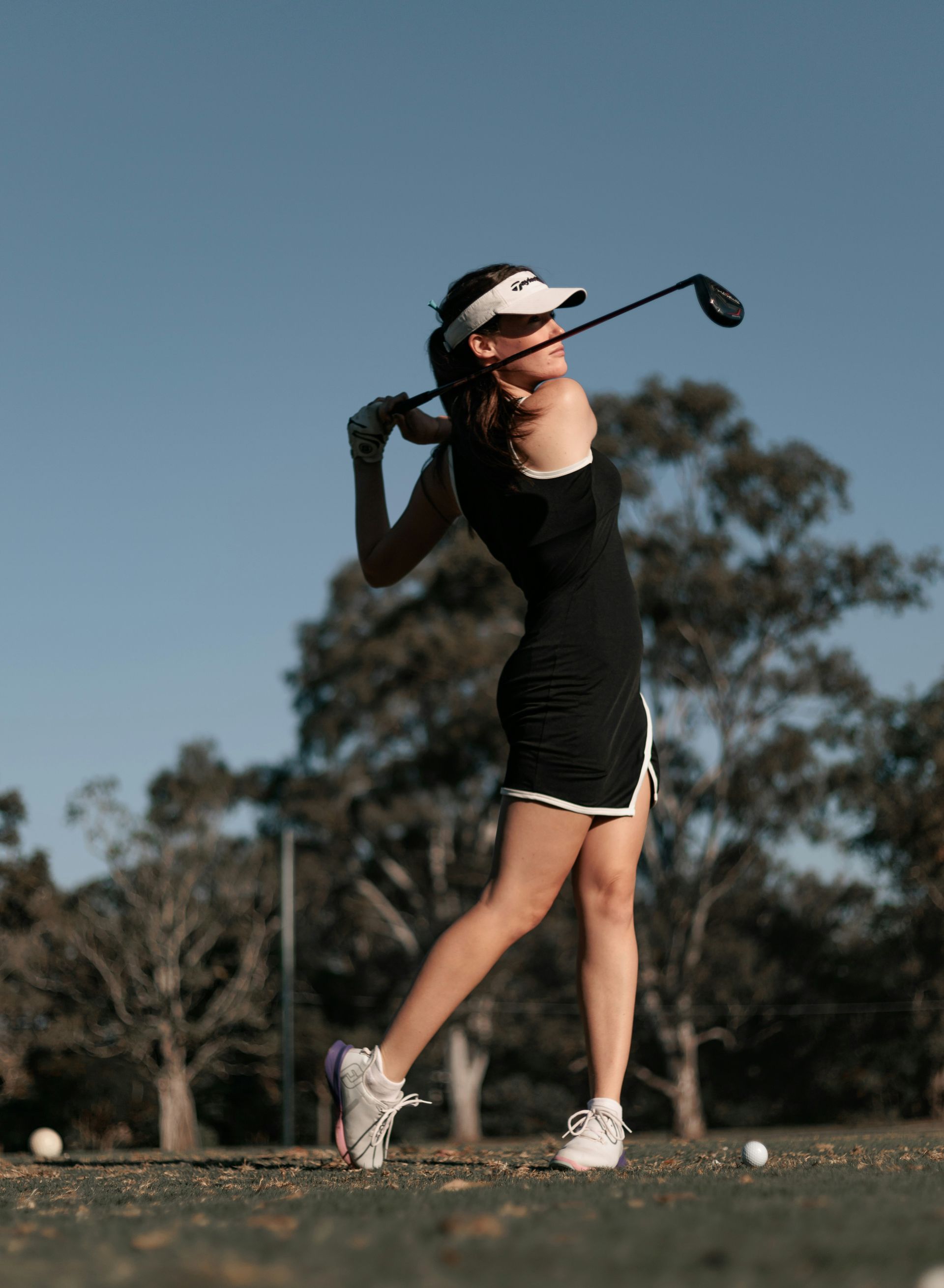 Woman in a black golf dress swings a club on a sunny day, trees in the background, with a golf ball nearby.