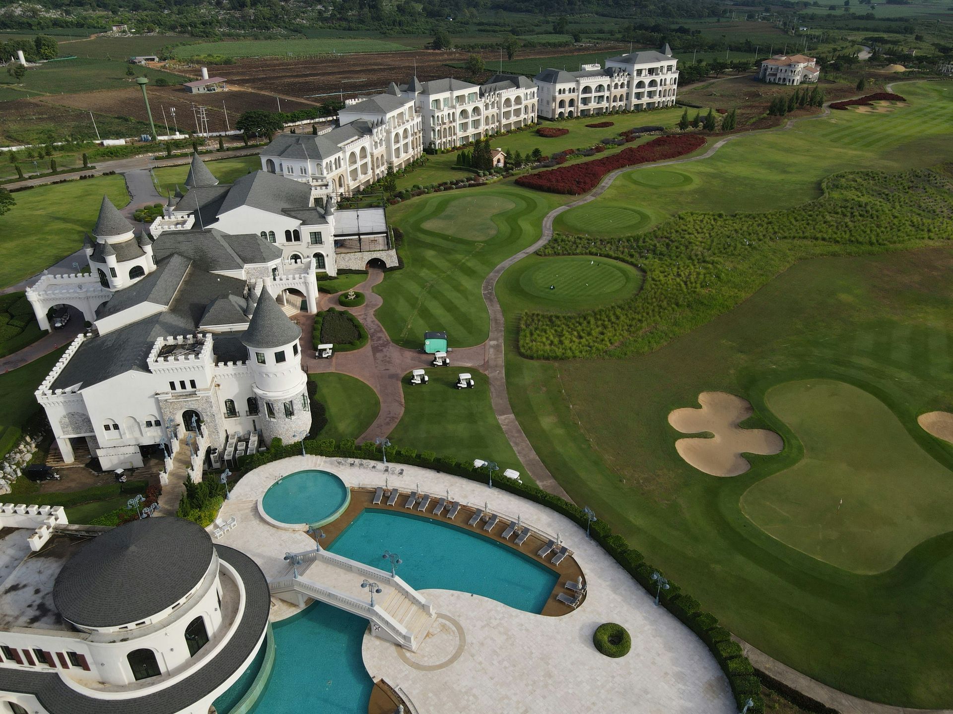 Aerial view of a white castle-like resort with a golf course, swimming pool, and apartment buildings. Green grass and blue water dominate the scene.