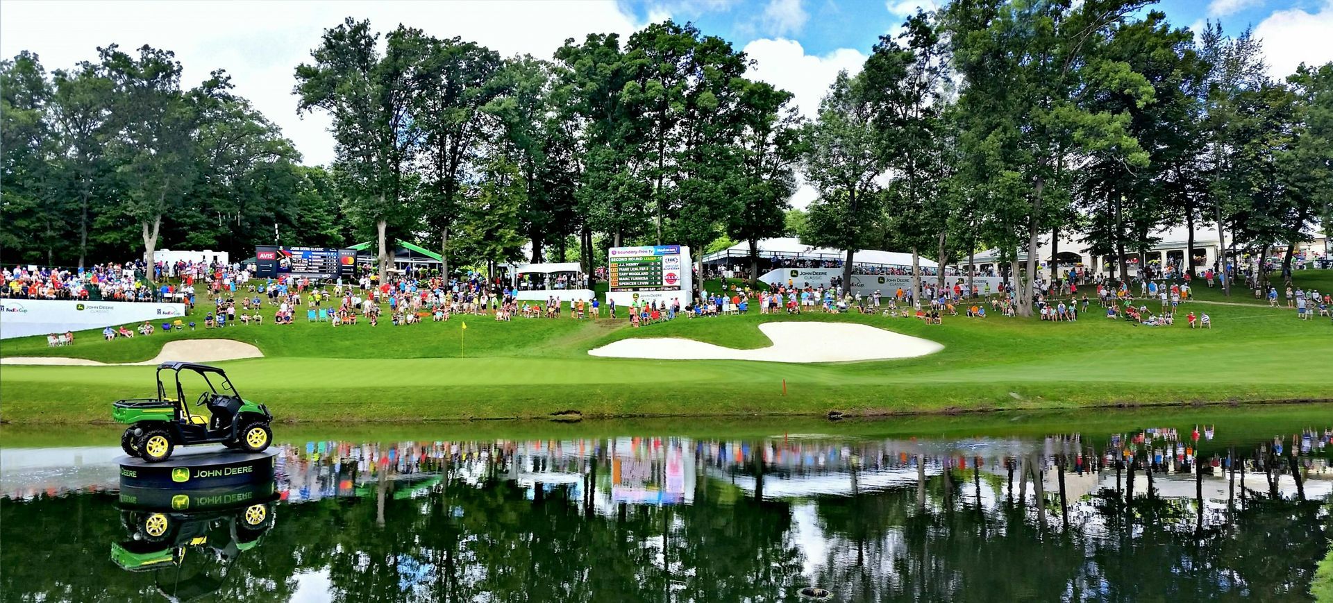 A golf course scene with spectators, trees, and a reflecting pond. A small green cart is in the water.