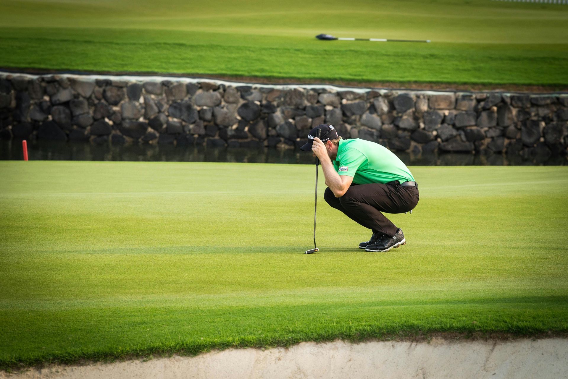 Golfer in green shirt and black pants lines up a putt on a green, with a water feature and green in the background.