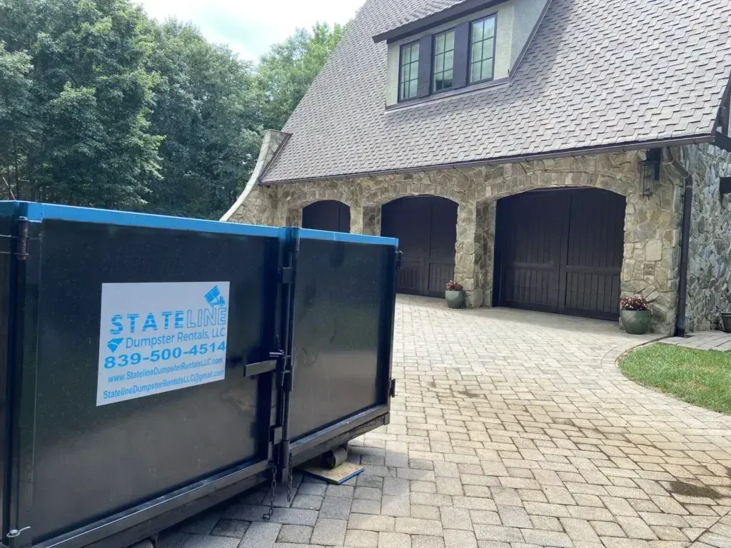 A large dumpster in front of a stone garage, driveway, and manicured lawn.