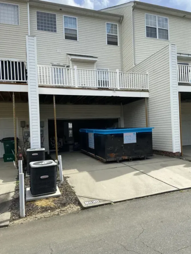 Townhouse exterior with a dumpster in the open garage beneath a deck; HVAC units on the left.