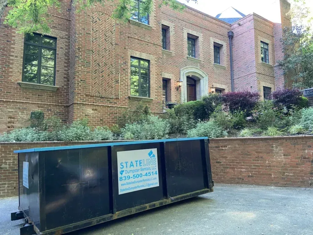 Black dumpster in front of a red brick building with windows and landscaping.