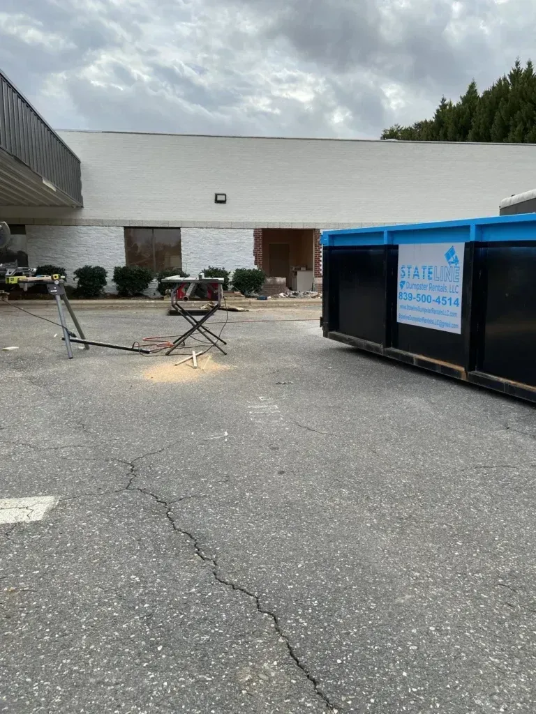 Construction site with a dumpster, saw, and debris. White building in the background. Asphalt parking lot. Overcast day.