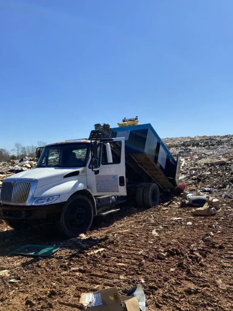 White garbage truck dumping waste at a landfill, blue sky overhead.