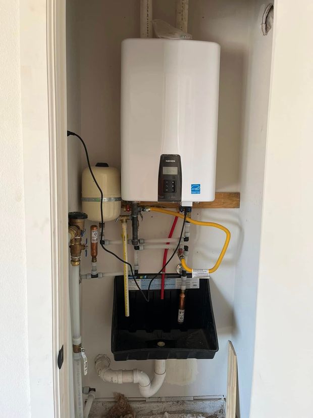 Wall-mounted white water heater in a utility closet with exposed pipes and hoses above a black sink basin