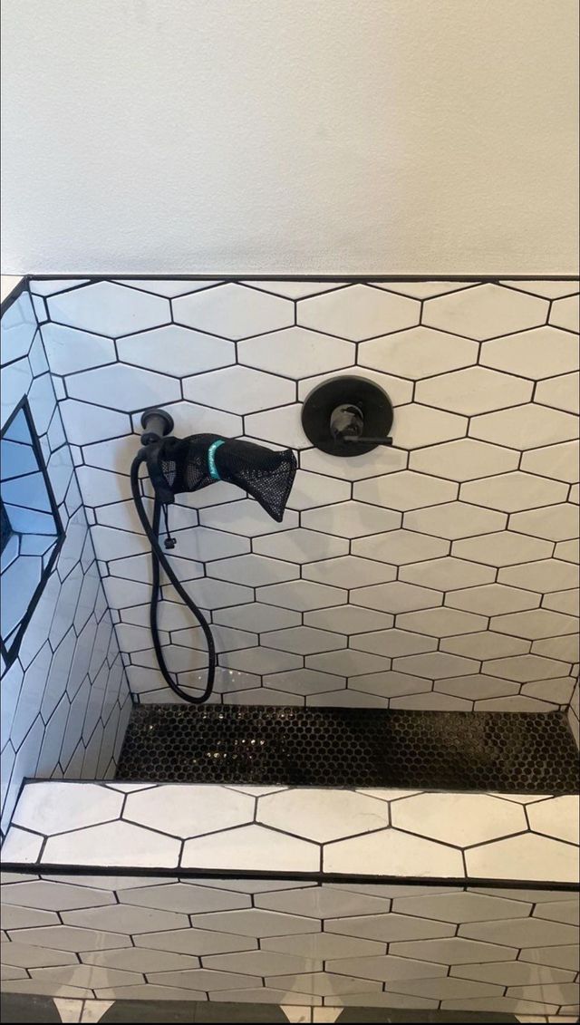White tiled shower wall with a black handheld showerhead and hose mounted beside a drain shelf.