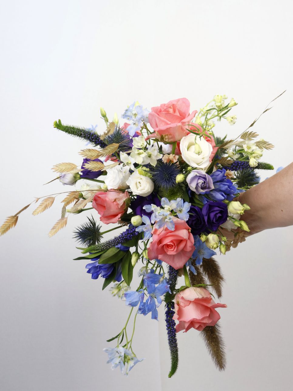 Een hand houdt een kleurrijk boeket vast met roze rozen, blauwe ridderspoor en witte bloemen.