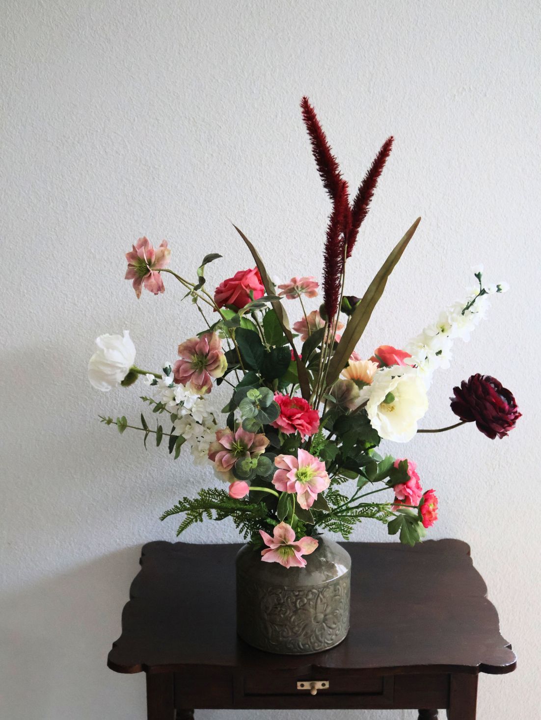 Zijdenloemen in een vaas op een donkere tafel tegen een witte muur; roze, rode en witte bloemen.