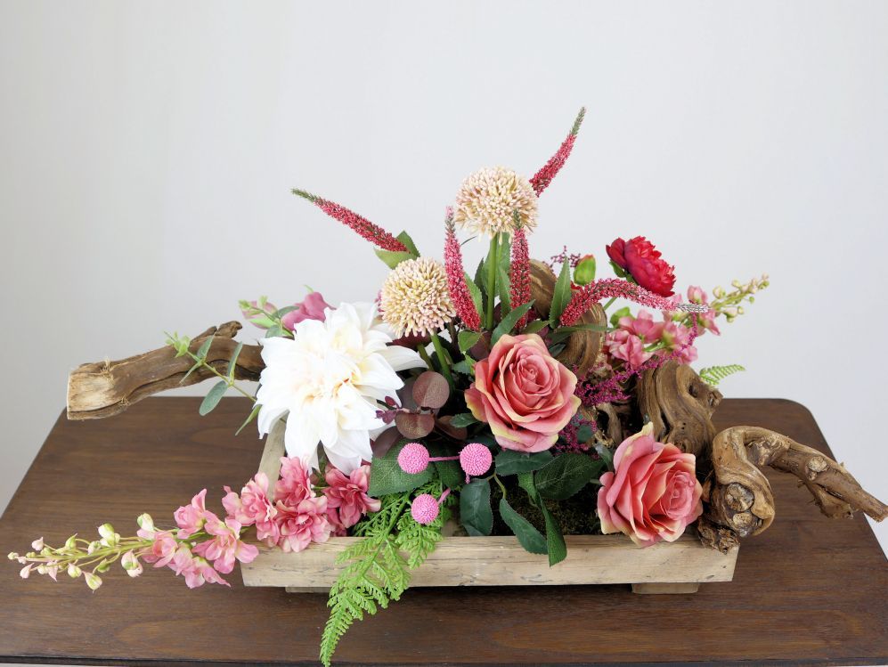 Bloemstuk in een houten schaal met zijdenbloemen rozen, dahlia's en groen met accenten van drijfhout.