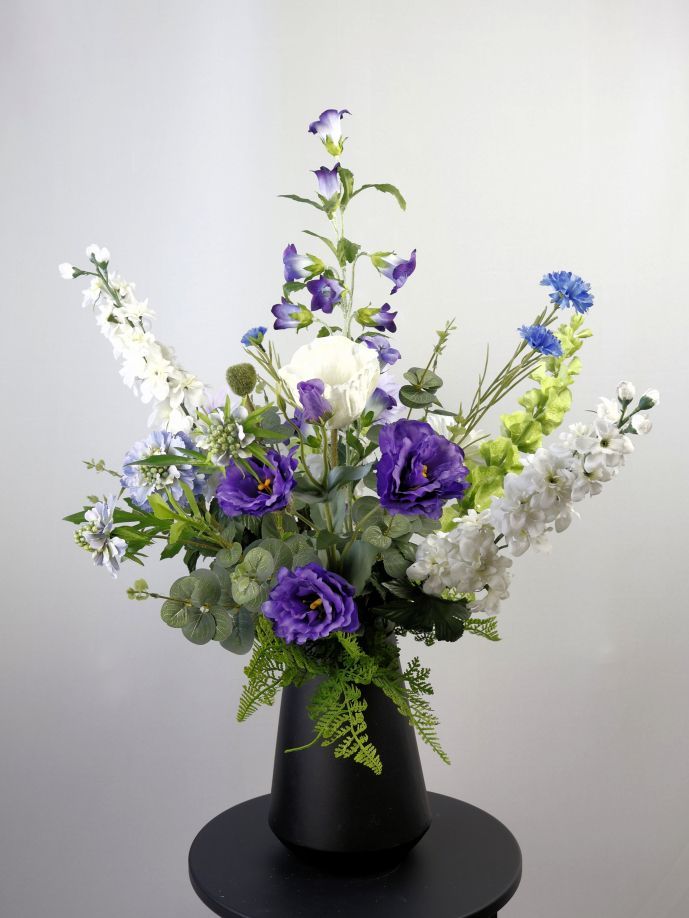 Een zijdenbloemen boeket met paarse, witte en blauwe bloemen in een zwarte vaas op een zwarte kruk.